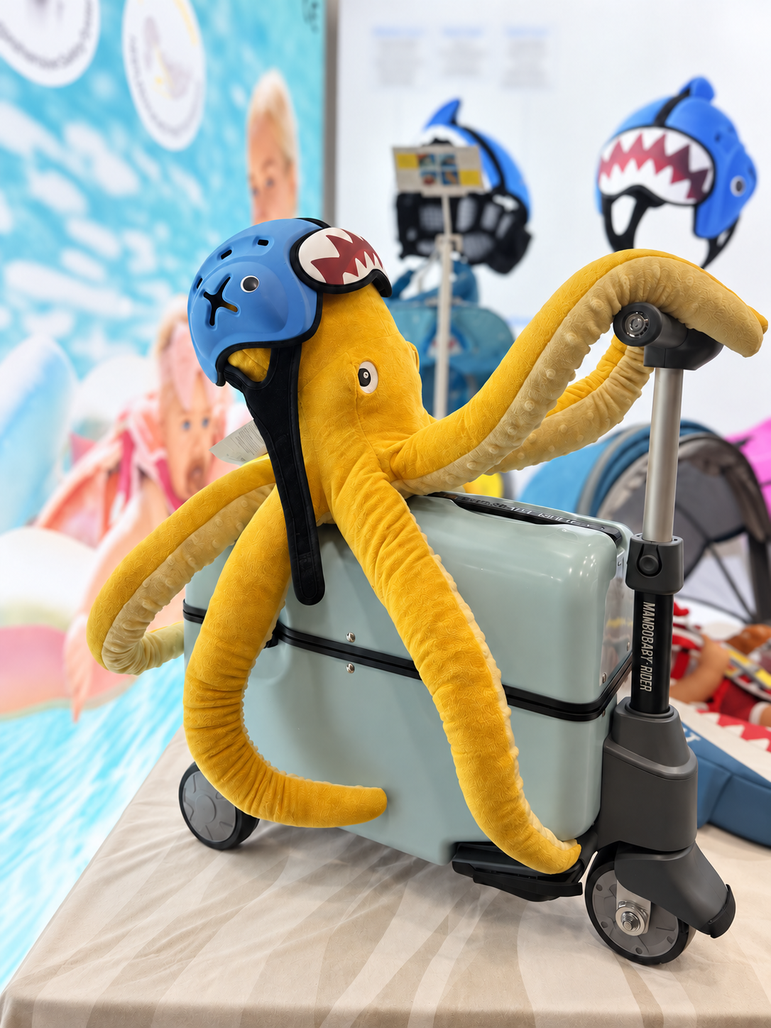Yellow octopus plush wearing a blue shark-themed Mambobaby Head Guard, seated on a gray-green Mambobaby Rider at the exhibition booth.