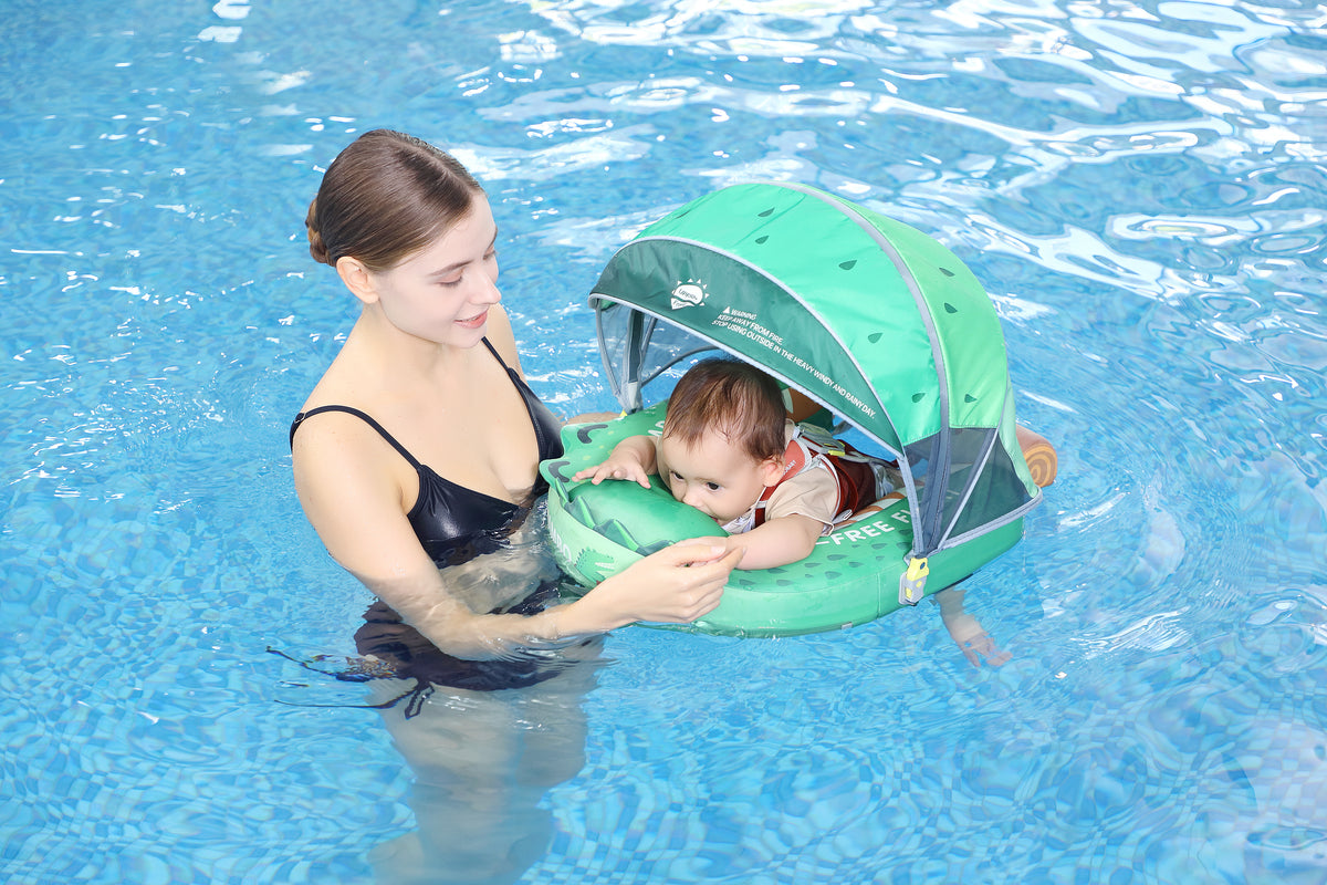 A baby wearing a beige outfit floats on a green water seat inside a swimming pool, with a woman in a black swimsuit holding them and a green canopy above the baby.